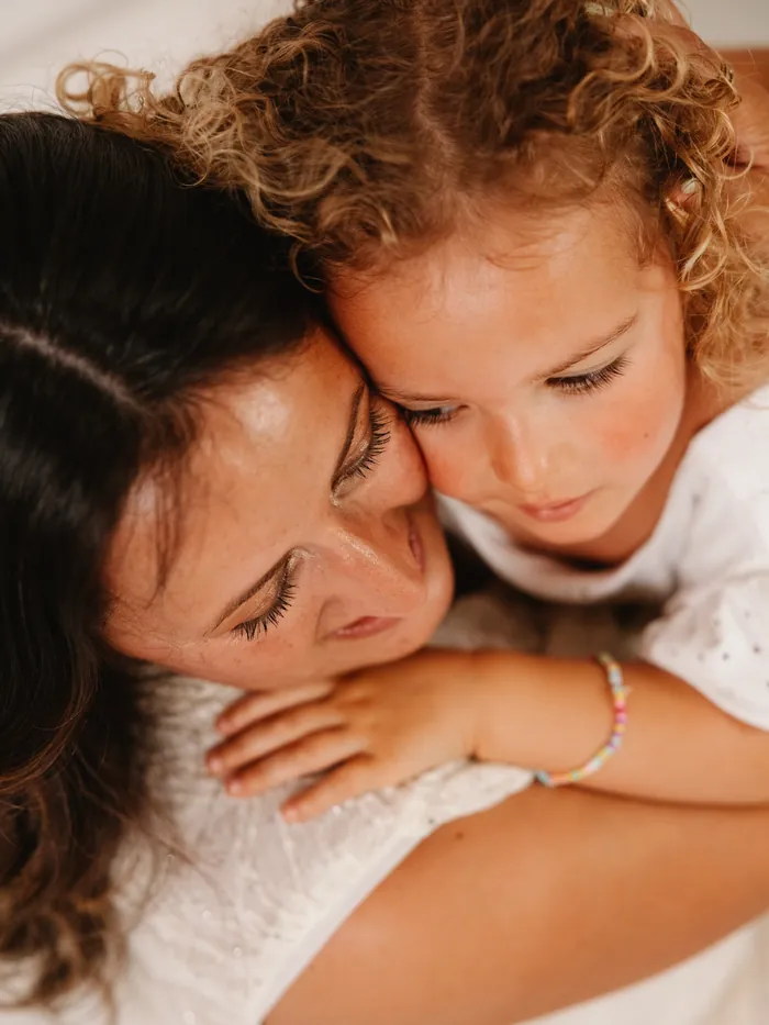 Mãe e filha abraçadas, cara com cara, a menina de caracóis loiros.