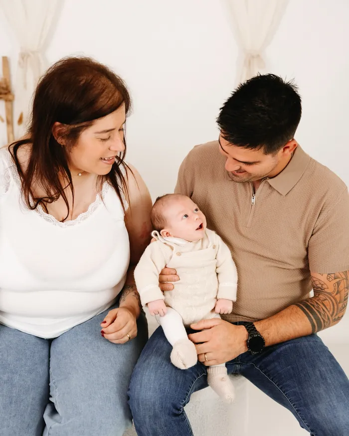 Pai e mãe sentados num banco de estúdio com o bebé de um mês ao colo.