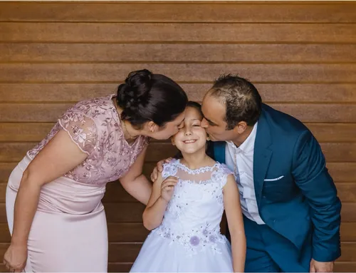 Menina com o vestido da comunhão, beijada pelo pai e pela mãe, à frente de uma parede de madeira.