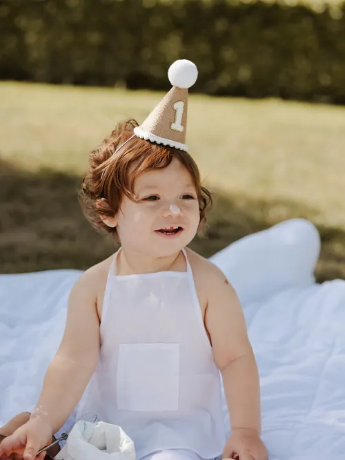 Bebé com um chapéu de festa com o número 1 e bolo no nariz, sentado numa manta na relva.