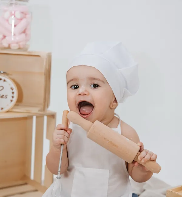 Bebé de avental e chapéu de cozinheiro, com um rolo da massa e uma vara de arames, num mesversário com tema.